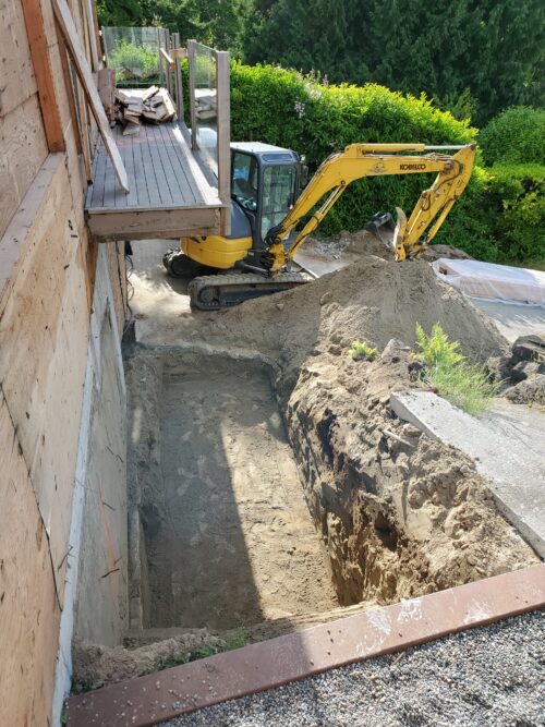 Construction photo of an excavator digging a foundation trench beside the North Beach home